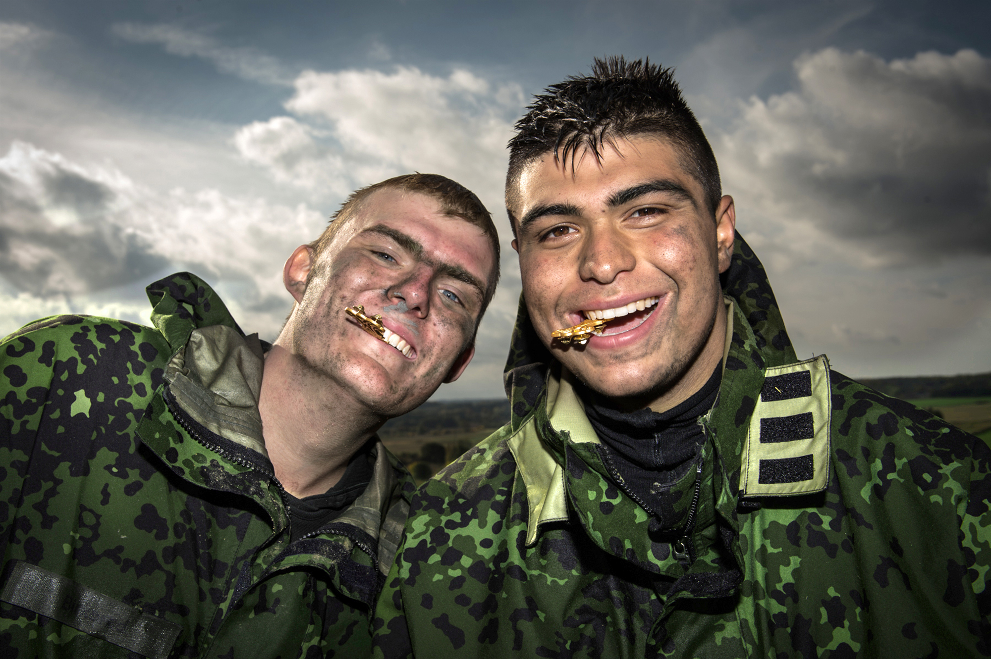 To unge mænd i camouflageuniformer med mudder i ansigterne smiler bredt, mens de hver især holder en guldfarvet genstand mellem tænderne; de står foran en overskyet himmel, hvilket giver billedet en rå stemning, og deres udtryk viser kammeratskab og glæde.