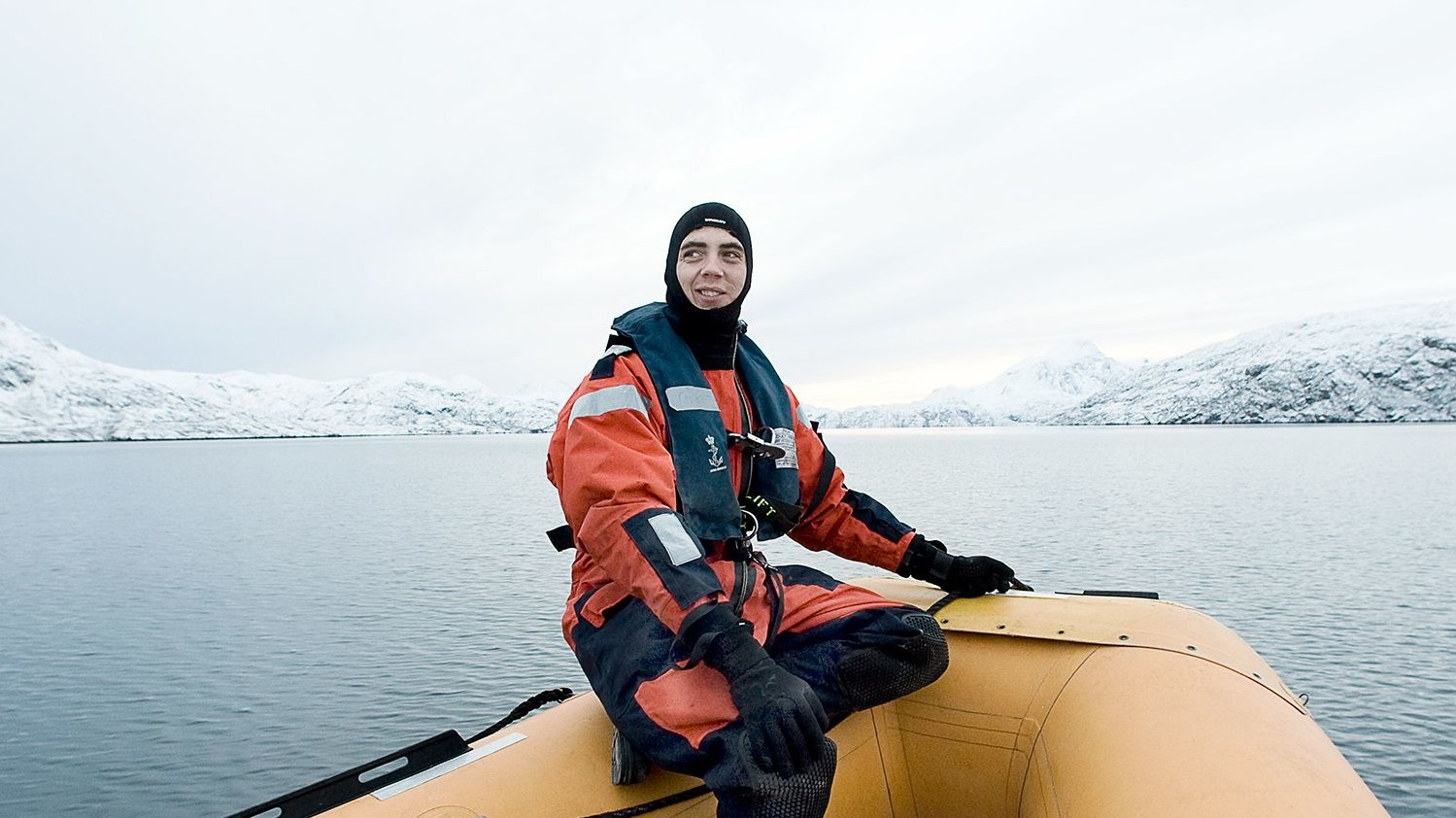 Person i redningsdragt sidder i en gummibåd på en rolig fjord omgivet af sneklædte bjerge.