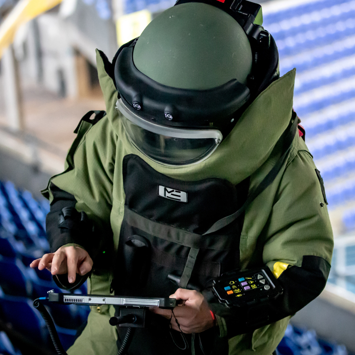 Person i bombedragts sikkerhedsudstyr bruger en håndholdt enhed i en tom sportsarena. Stemningen er alvorlig.