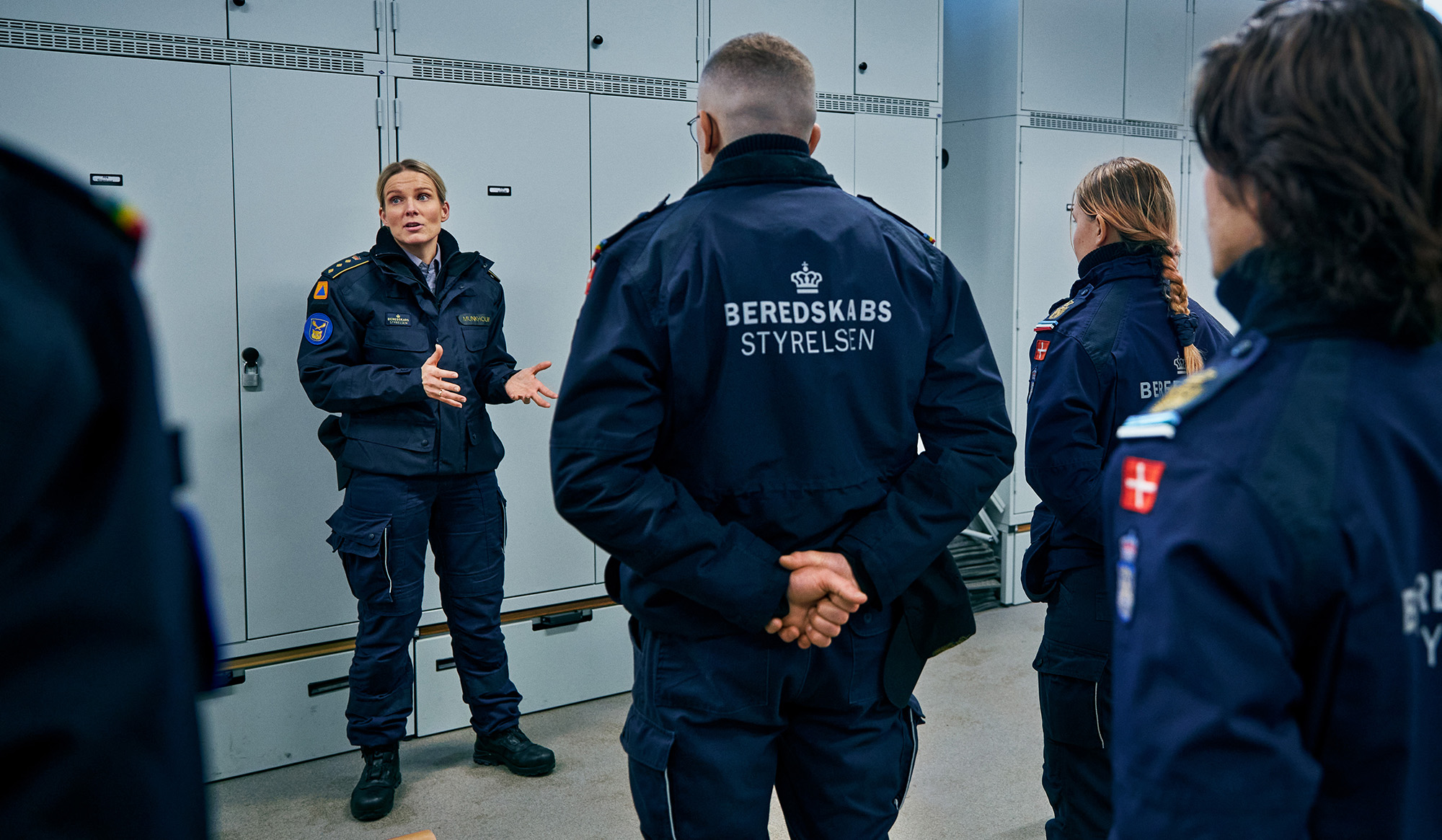 En kvinde i uniform, sandsynligvis en leder eller instruktør, taler engageret til en gruppe personer i ens uniformer under en briefing eller træning, hvor hun bruger håndbevægelser for at understrege sine pointer; i baggrunden ses store skabe, hvilket antyder et struktureret miljø relateret til beredskab eller sikkerhedstjenester, og uniformerne har insignier og logoer, der viser tilhørsforhold til en bestemt organisation.