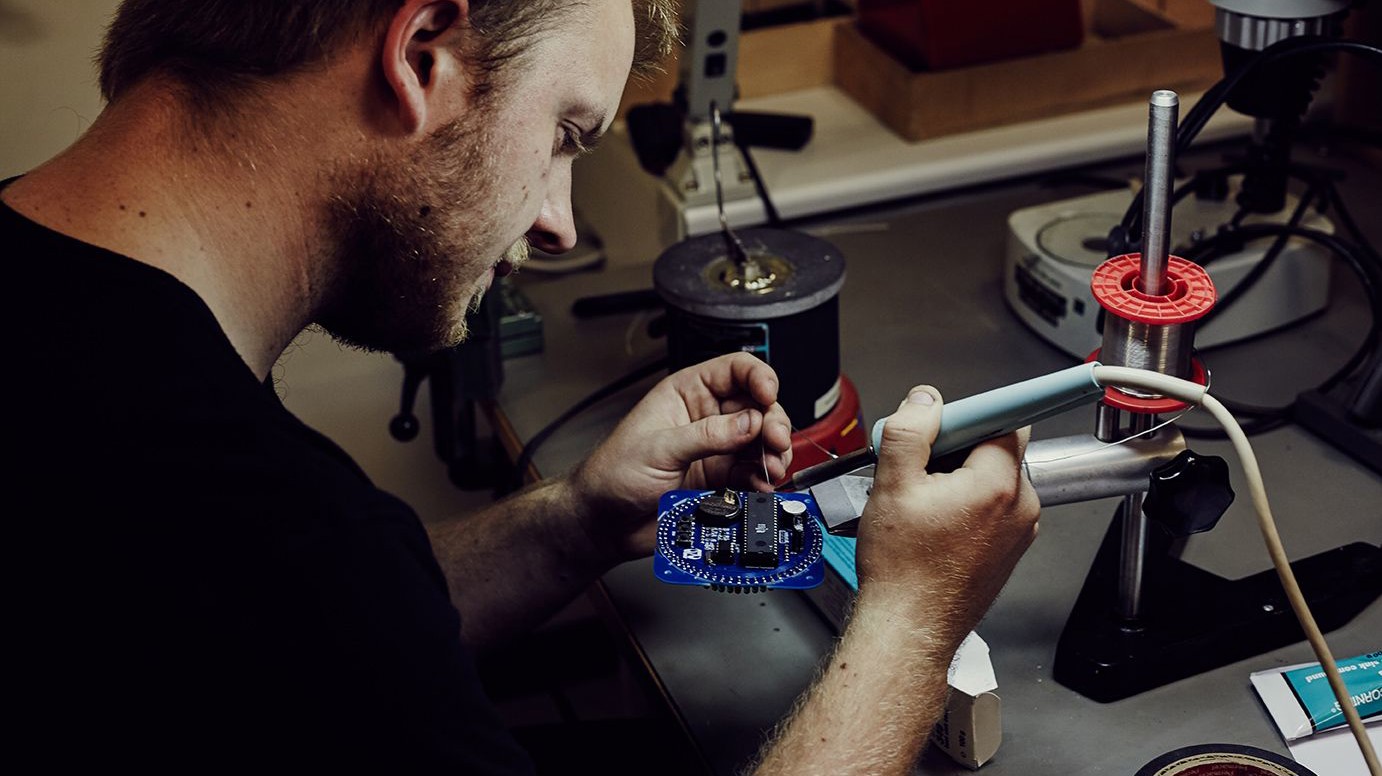 En person arbejder koncentreret med at lodde elektroniske komponenter på et printkort med loddekolbe og fin tråd.