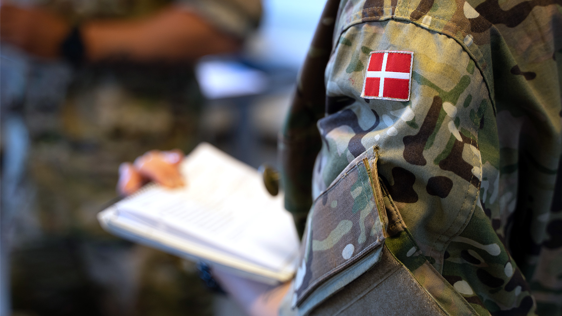 Soldat i camouflageuniform med dansk flagmærke på skulderen, en hånd holder en notesblok i forgrunden, sløret militær baggrund.
