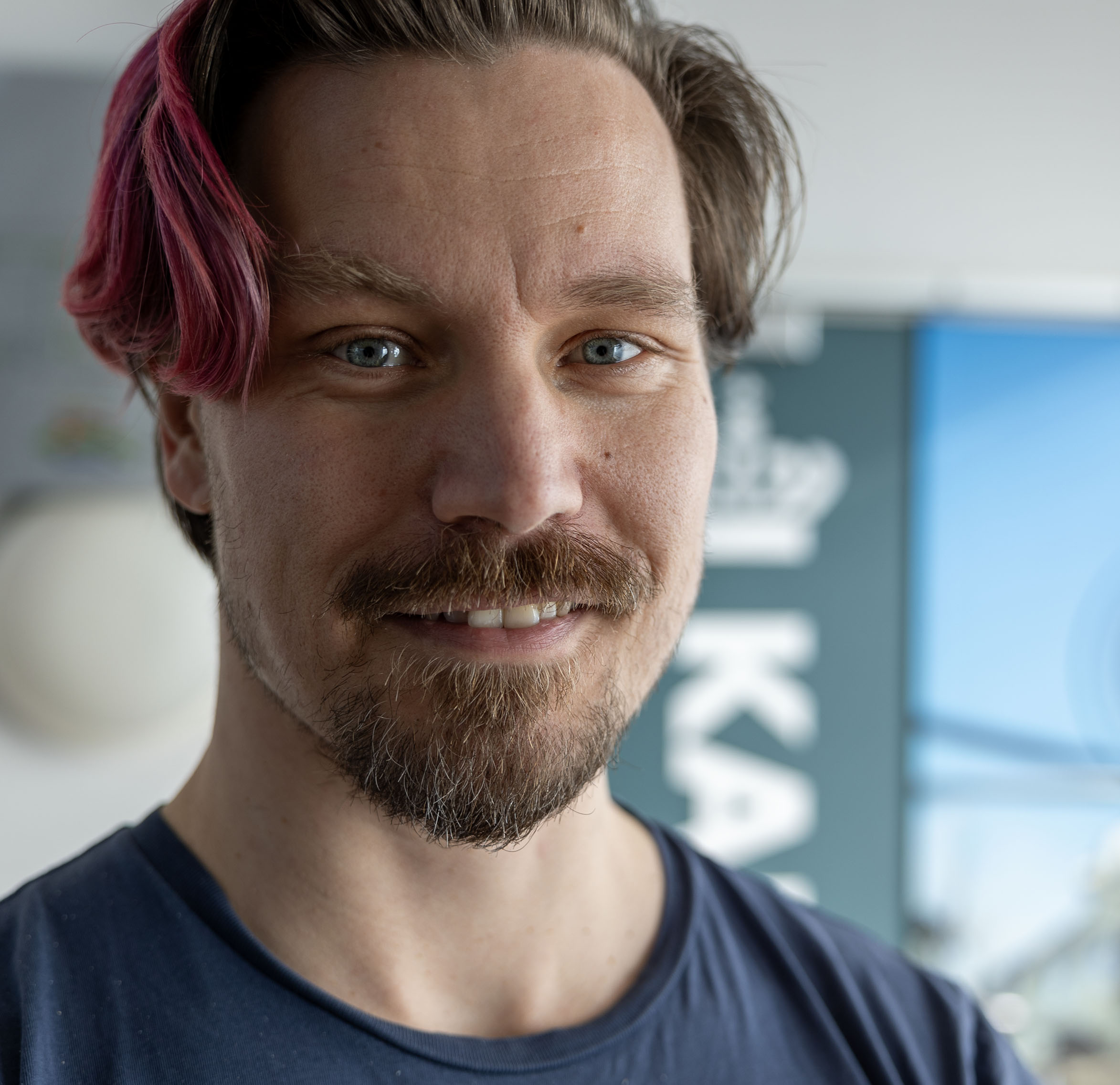 Mand med lysebrunt og pink hår, velplejet skæg og blå øjne i navyblå t-shirt med sløret baggrund.