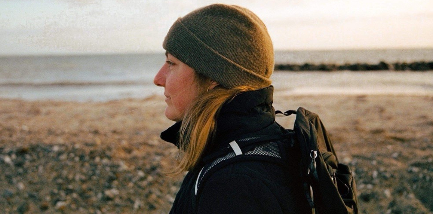 Kvinde med langt hår i brun hue og mørk jakke kigger mod højre ved kysten i blødt solnedgangslys, bærer rygsæk.