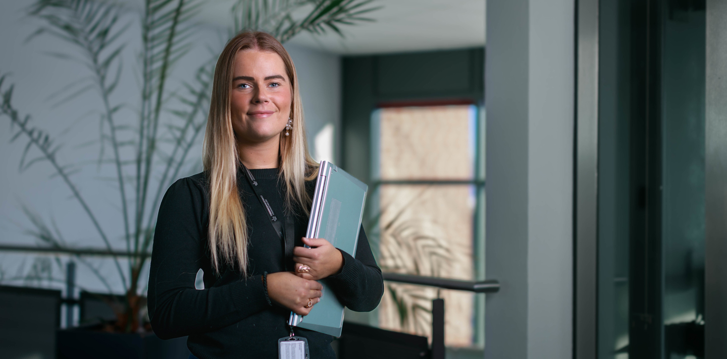 Ung kvinde i sort top står selvsikkert indendørs med mapper i hånden og grønne planter og store vinduer i baggrunden.