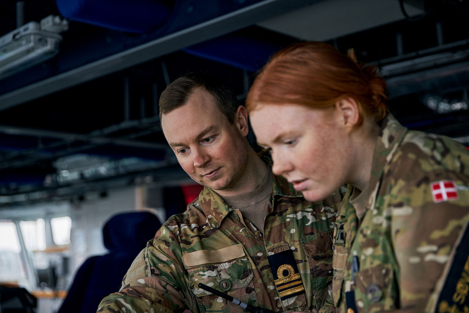 To militærpersoner arbejder sammen iført camouflageuniformer. Kvinden til højre bærer et særpræget mærke med det danske flag. De befinder sig i et skibsinteriør, muligvis et kommandorum, hvor teknisk udstyr ses i baggrunden. Begge har et koncentreret udtryk, hvilket antyder samarbejde om en teknisk eller operativ opgave.