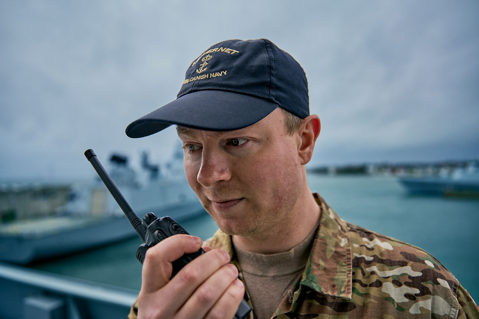 Billedet viser en mandlig militærperson i camouflageuniform og en mørkeblå kasket med teksten "Royal Danish Navy." Han er koncentreret om en håndholdt radio, hvilket indikerer en kommunikationssituation. I baggrunden ses flådefartøjer og en overskyet himmel, hvilket antyder et maritimt miljø. Personens udtryk udstråler koncentration og alvor, mens han udfører kommunikationsopgaver.