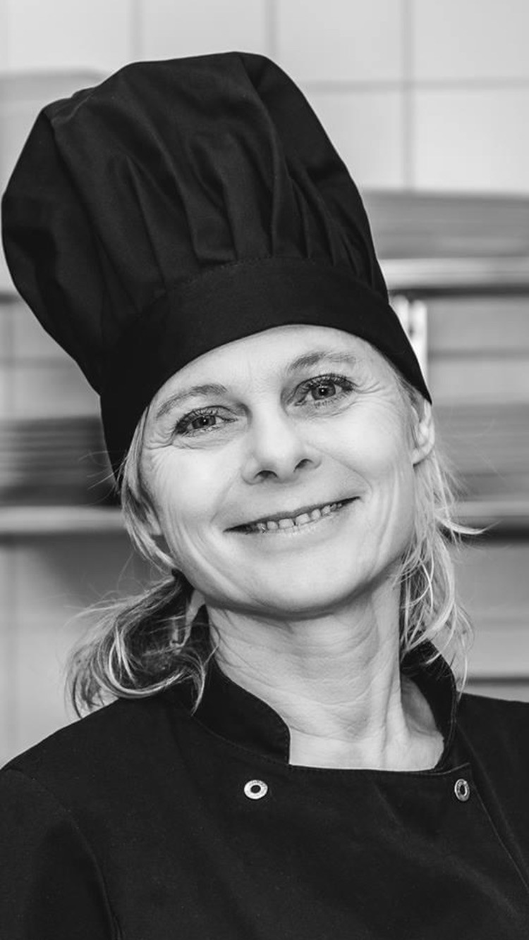 Kvinde i sort kokkehue og jakke smiler i et køkkenmiljø. Sort-hvid billede fremhæver hendes glade udtryk og professionelle tøj.