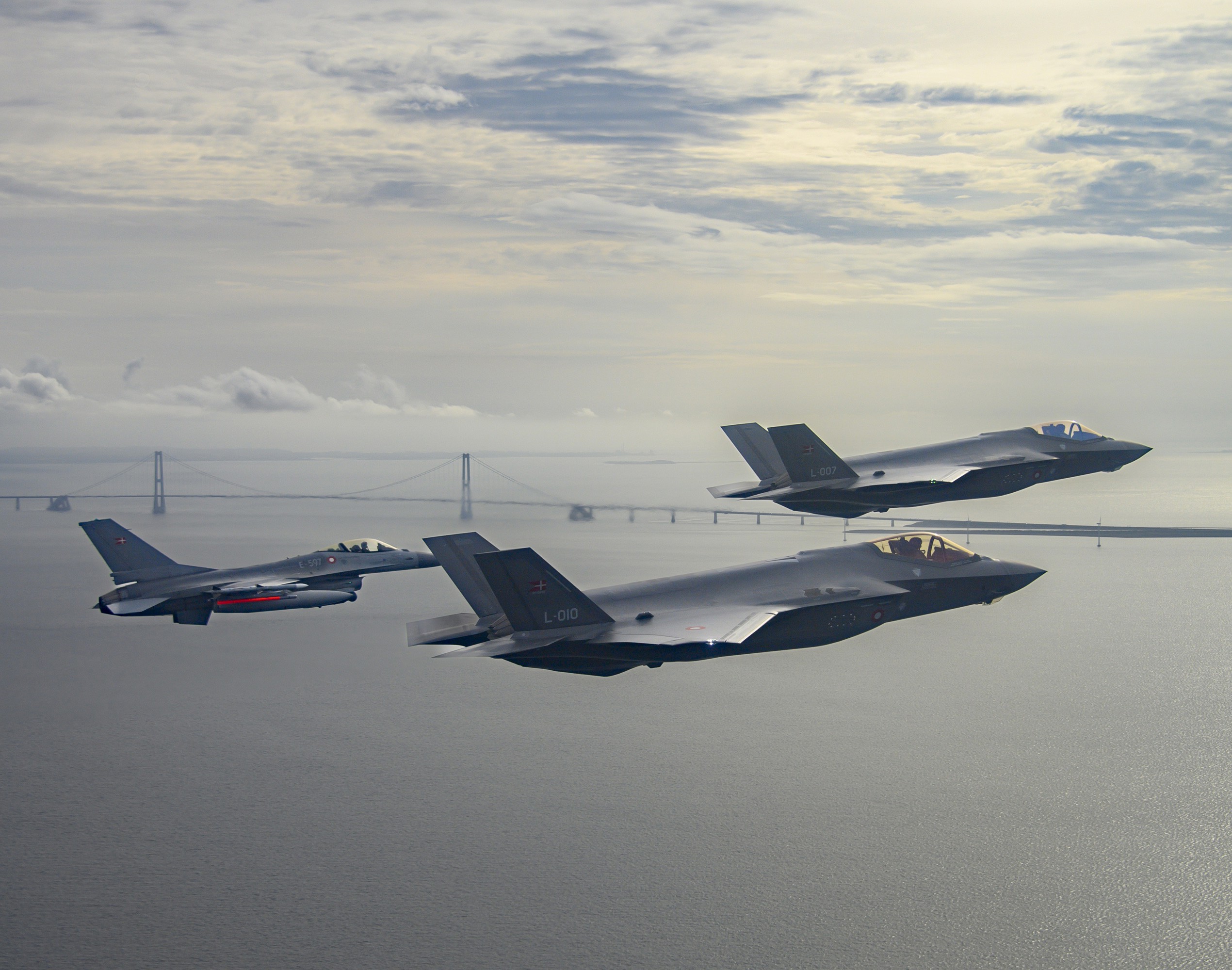 Tre militærfly flyver i formation over vand med en bro i baggrunden under en dramatisk, overskyet himmel.