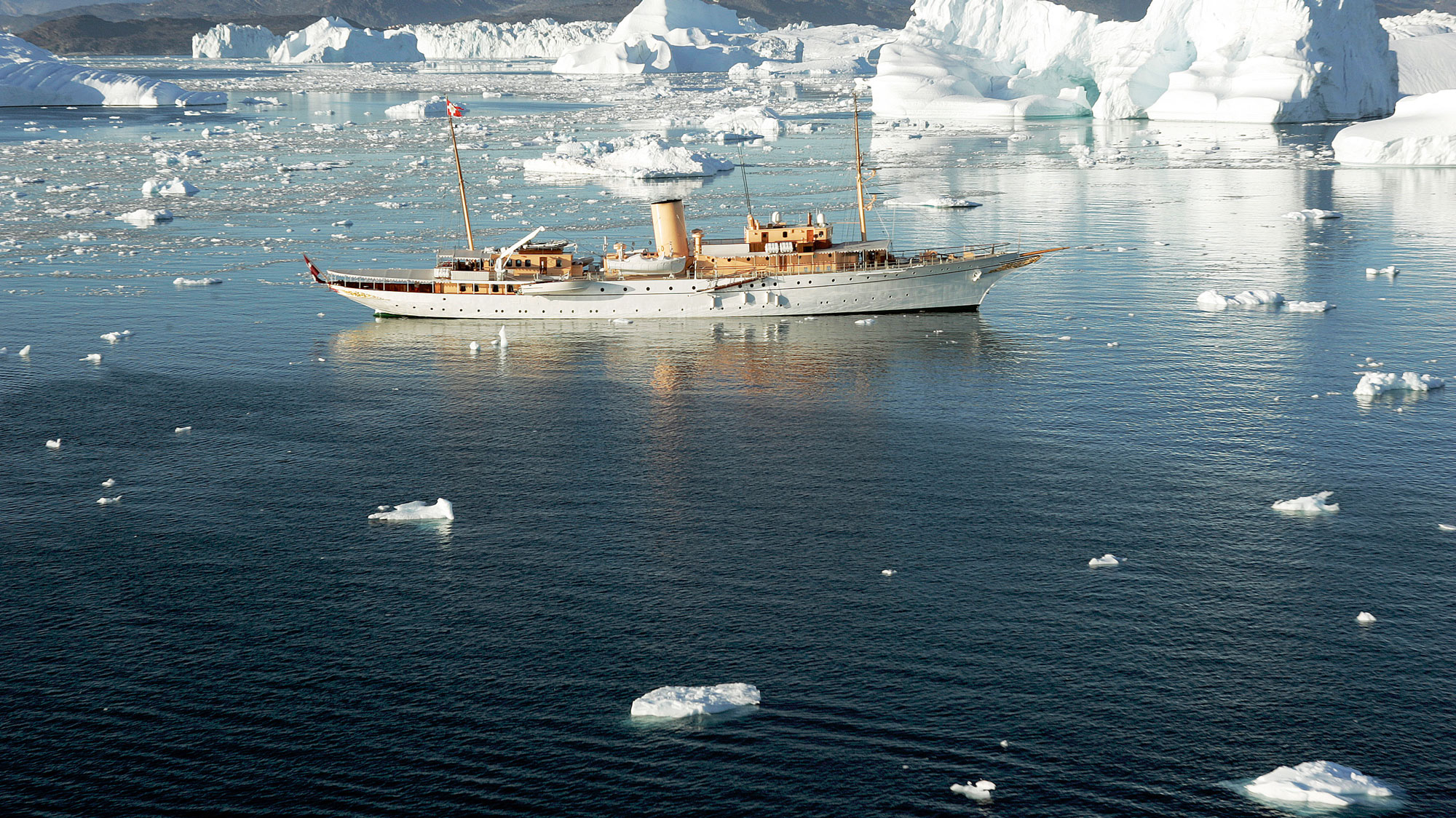 Klassisk yacht med trædetaljer og to master sejler mellem isbjerge i klart, spejlende vand i et roligt, polart landskab.