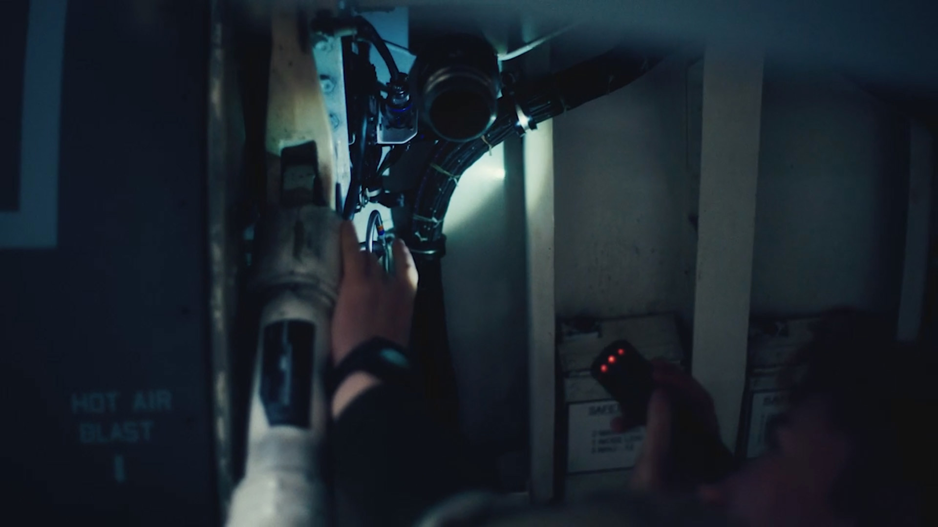 En hånd rækker mod et teknisk komponent med knapper og ledninger i svagt blåt lys, der antyder vedligeholdelse i trange omgivelser.