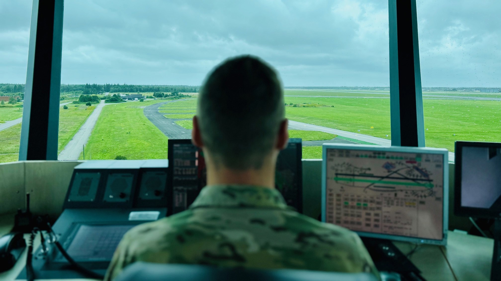 Militærperson sidder i kontroltårn og overvåger landingsbane og grønne marker via flere computerskærme under overskyet himmel.