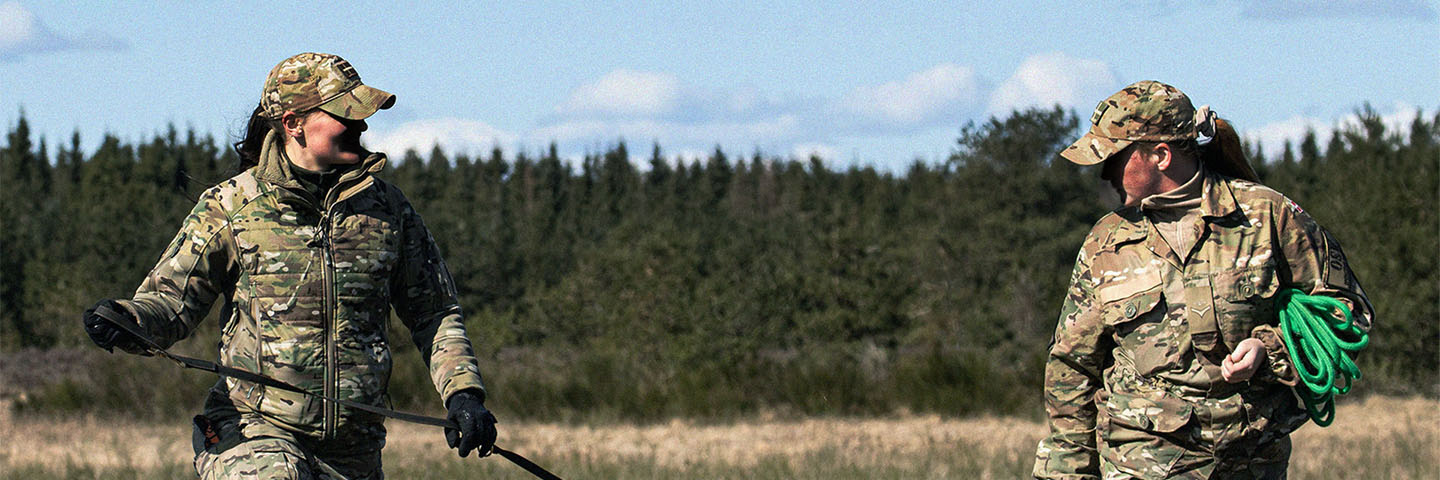 To personer i camouflageuniformer går med en sort hund med orange bold i munden i et græsbevokset, udendørs område.
