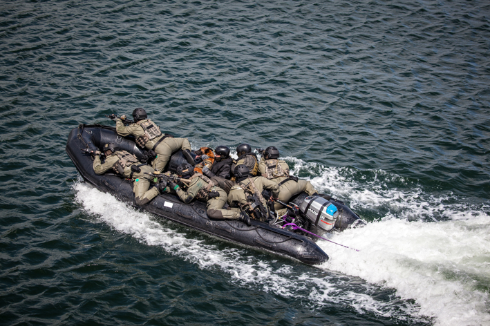 En gruppe personer i taktisk udstyr på en sort gummibåd bevæger sig hurtigt gennem vandet, koncentrerede og målrettede.
