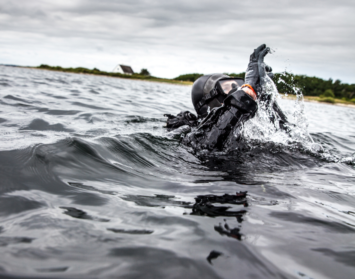 En dykker i sort våddragt stikker op af vandet og laver en håndbevægelse, med skyer og en hvid bygning i baggrunden.