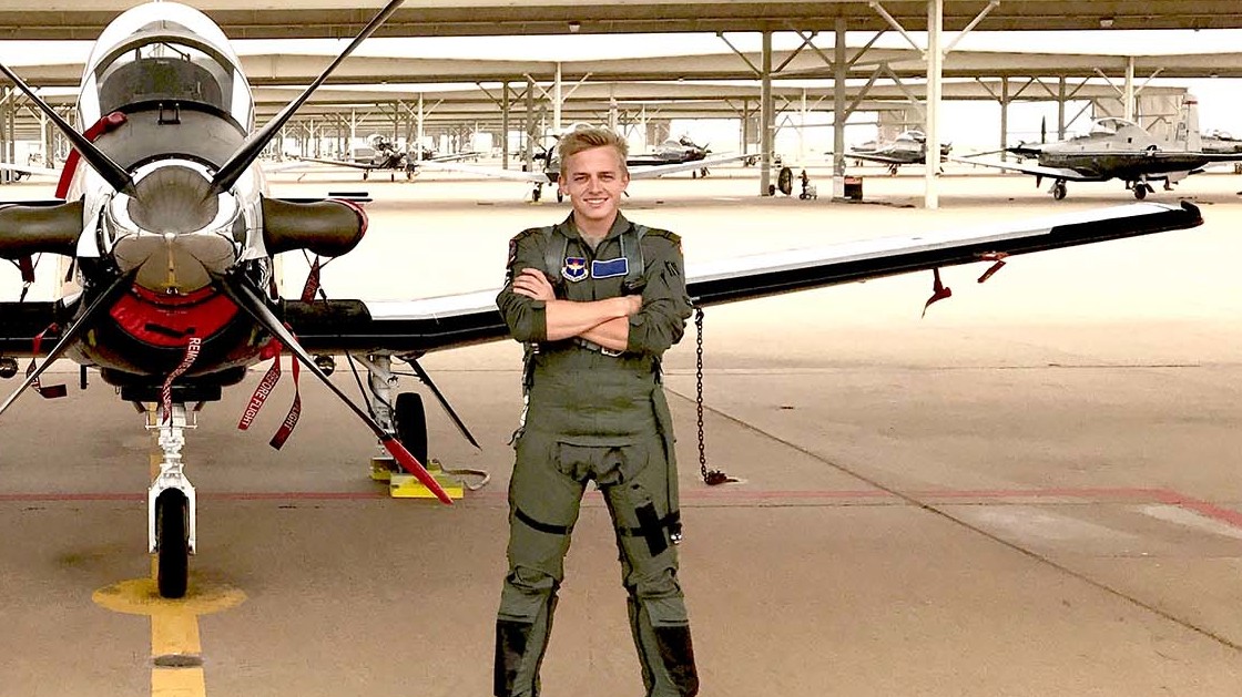 Ung pilot i flyverdragt står selvsikkert foran et T-6 Texan II militærfly med korslagte arme på en aktiv flyveplads.
