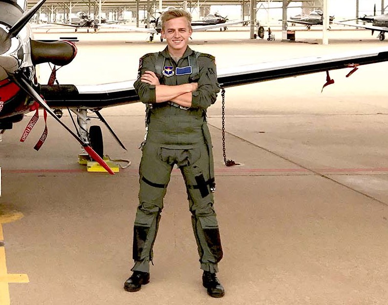 Person i militær flyverdragt står med korslagte arme foran et fly og smiler, omgivet af flere fly i en hangar.