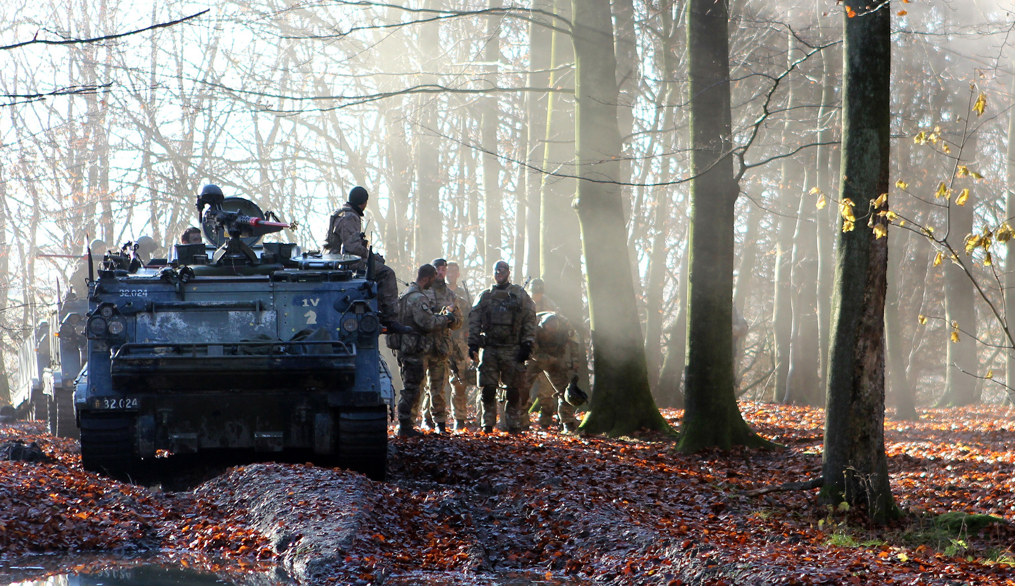 Soldater i camouflage uniformer samles ved et pansret køretøj i en skov med tåge og nedfaldne blade, der skaber en stemningsfuld scene.