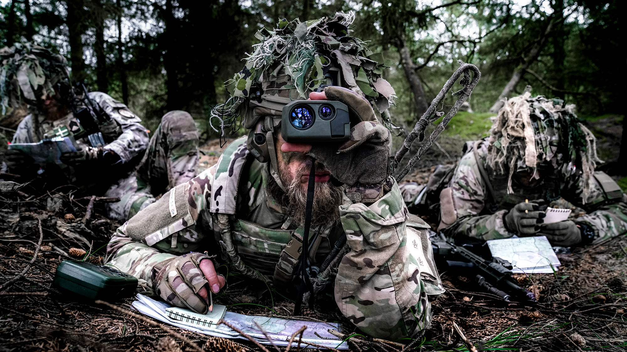 Soldat i camouflagetøj og ghillie suit bruger kikkert i skov, mens kollegaer arbejder med kort og notesbog. Fokus på rekognoscering.