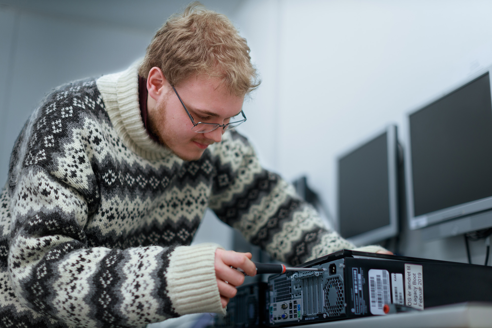 Ung mand i mønstret sweater arbejder koncentreret på at reparere en computertårn i et kontor- eller laboratoriemiljø.