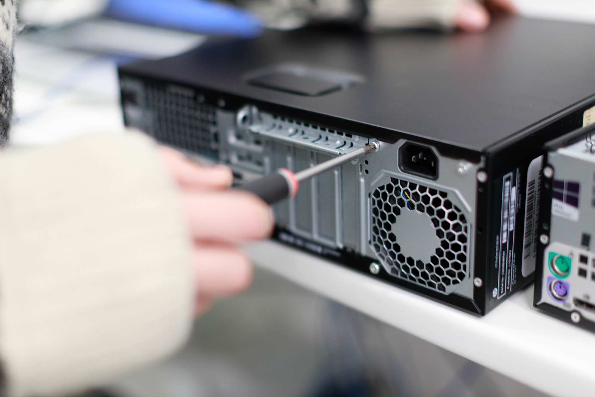 En person i lys sweater bruger en skruetrækker på bagsiden af et sort computerkabinet med ventilation og porte.