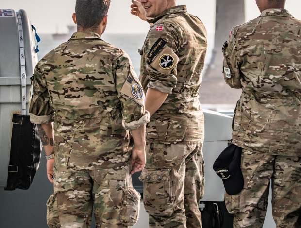 Tre soldater i camouflageuniform taler sammen. På den ene er det danske flag og Søværnets mærke synligt på ærmet.