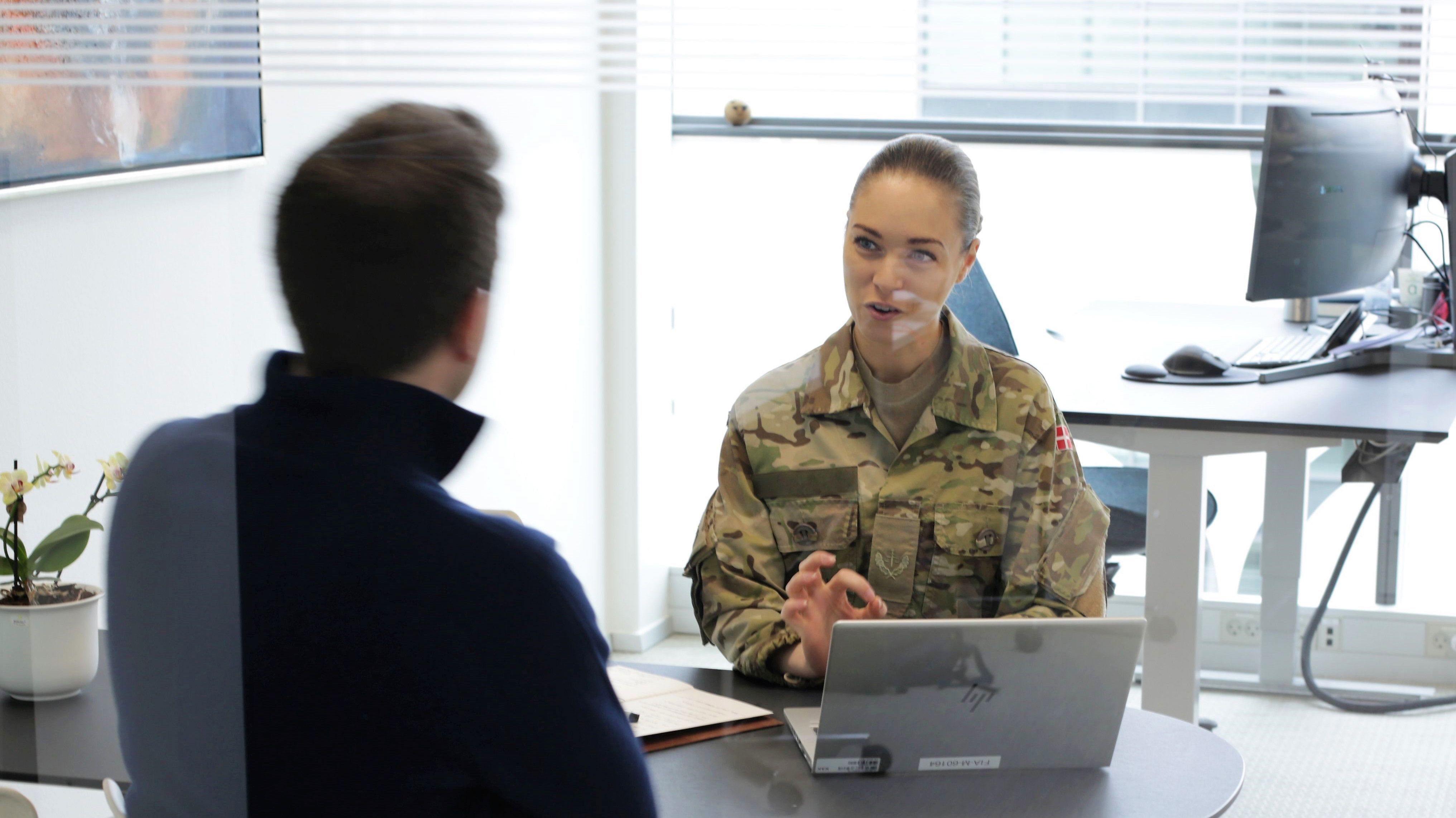 Soldat i uniform taler med en civil mand ved et bord under en samtale på kontor.