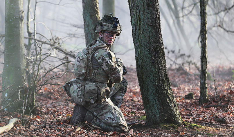 Soldat sidder i skoven i fuld uniform. Han kigger mod kameraet.