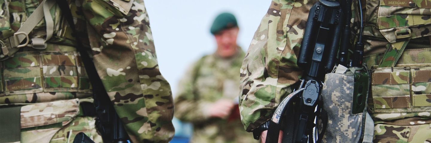 Ryggen af to kampklædte soldater. Foran dem strå en befalingsmand som er sløret.