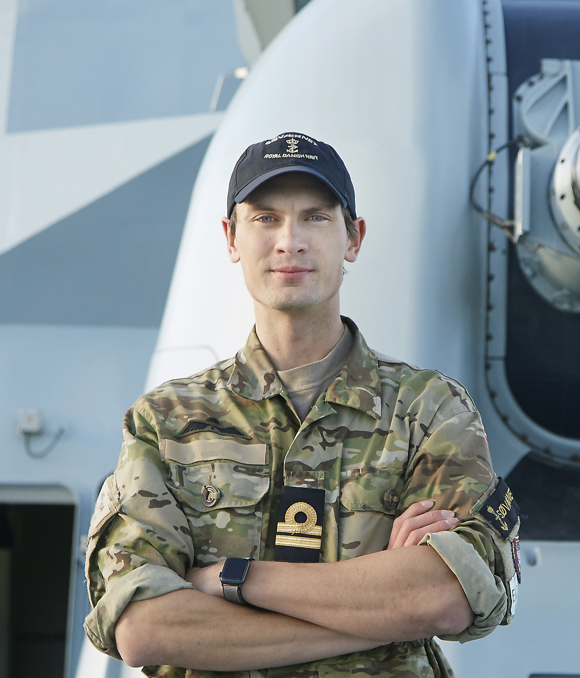 Militærperson iført camouflageuniform står selvsikkert med korslagte arme. Personen bærer en marineblå kasket med insignier, der angiver tilknytning til flåden. Uniformen har militærmærker på ærmerne samt en gylden ranginsignie. Baggrunden antyder et maritimt miljø, sandsynligvis ombord på et skib, med synligt maskineri.