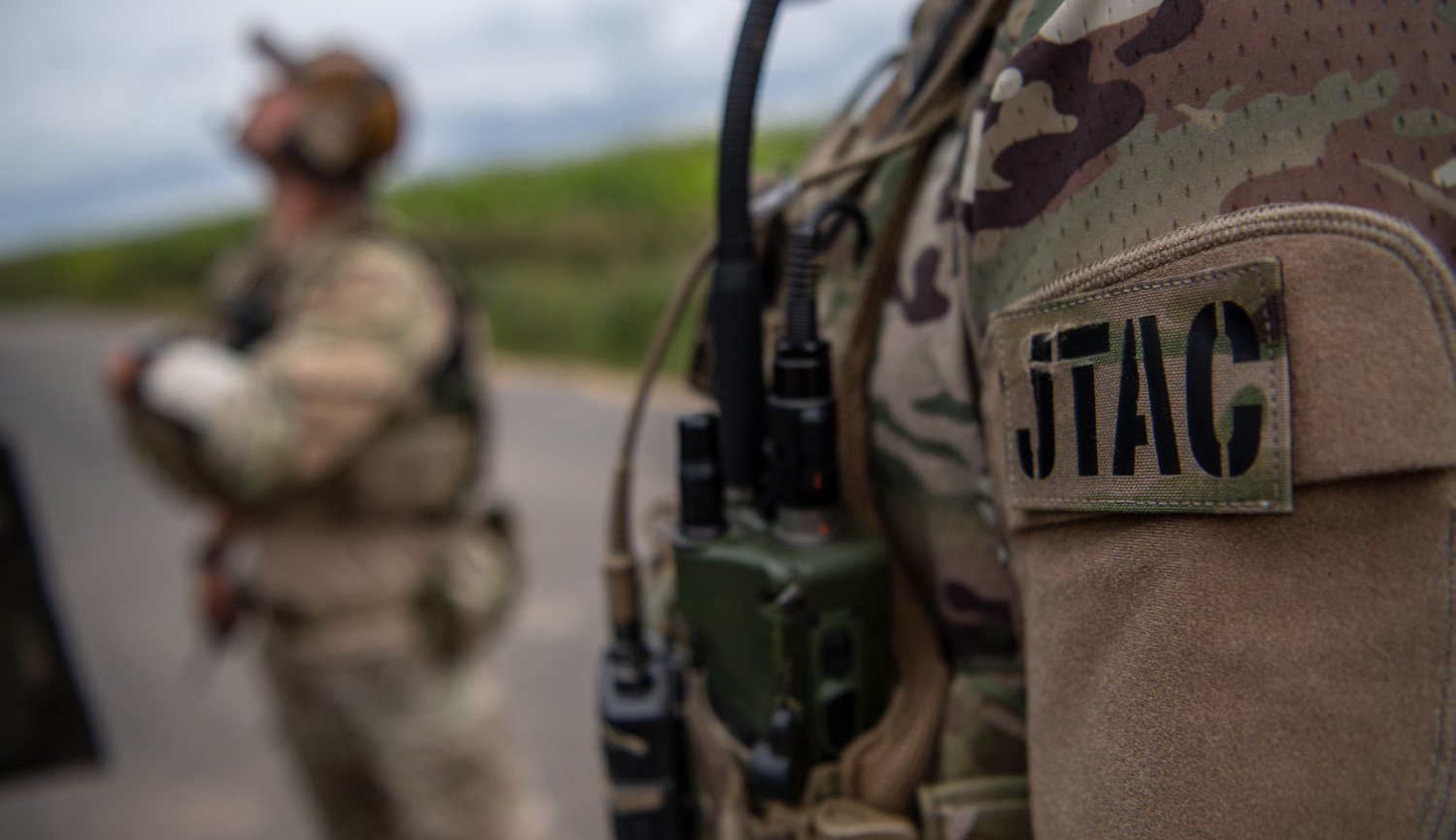Soldat i camouflageuniform med JTAC-patch og kommunikationsudstyr, står udendørs med sløret kollega og grøn baggrund.