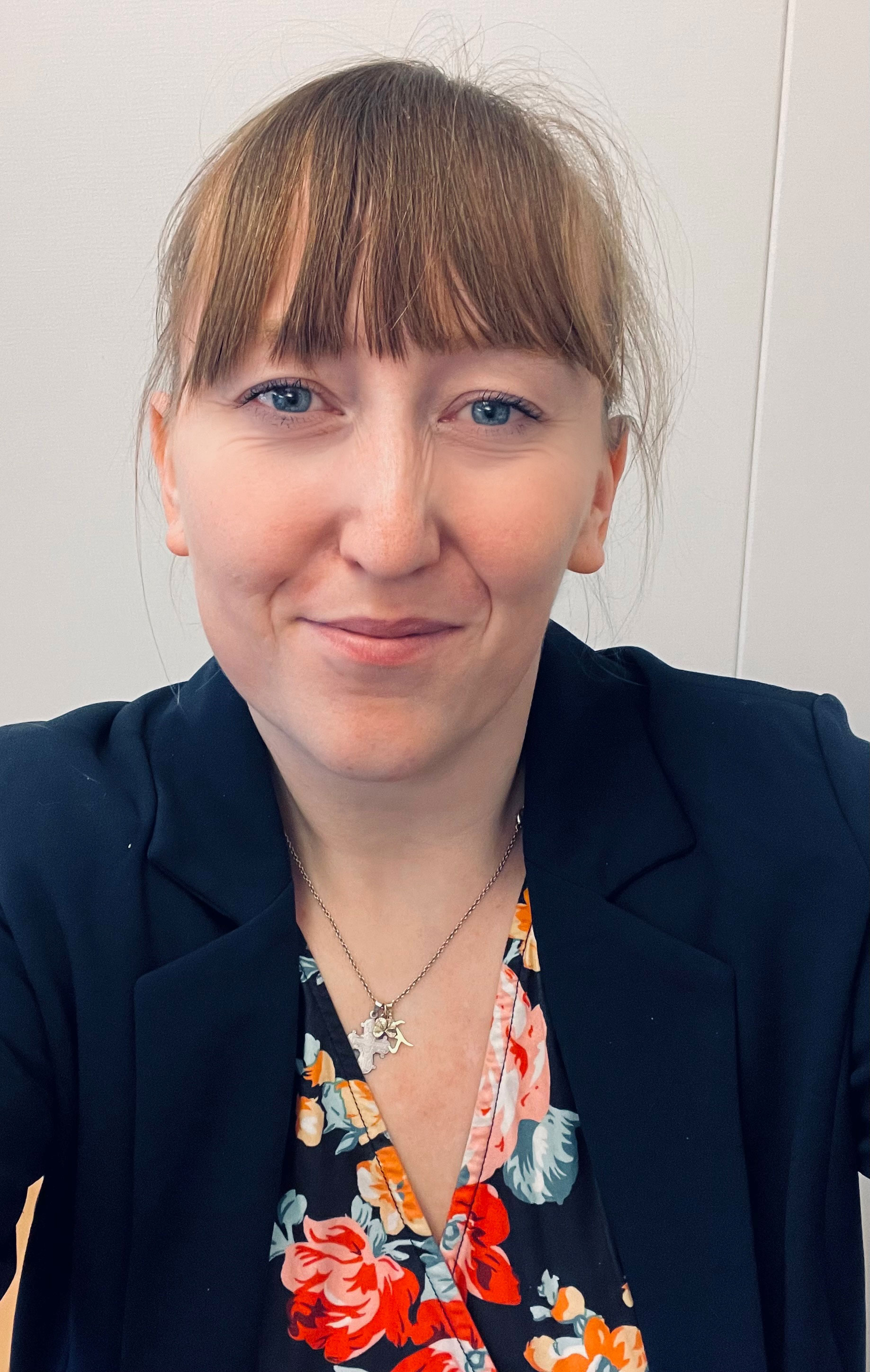 Billedet viser en kvinde med lysebrunt hår med pandehår og blå øjne. Hun har en mørk blazer på over en blomstret top i livlige farver, og hun bærer en sølvhalskæde med et vedhæng. Baggrunden er ensfarvet og neutral, hvilket fremhæver hendes venlige og imødekommende udtryk.