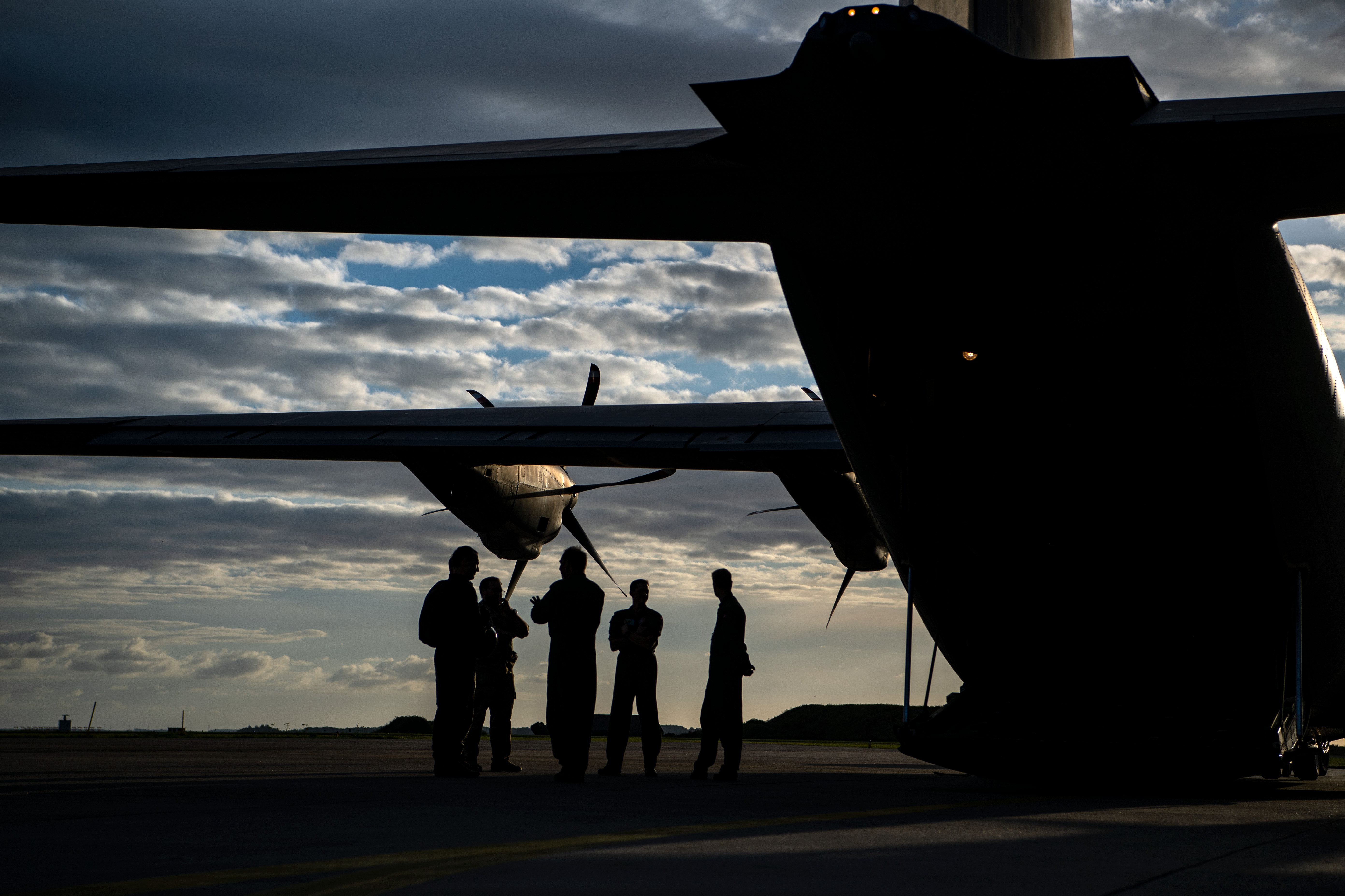 Silhuetter af personer står under et flys vinger på en flyveplads i aftensol, omgivet af dramatiske skyer.