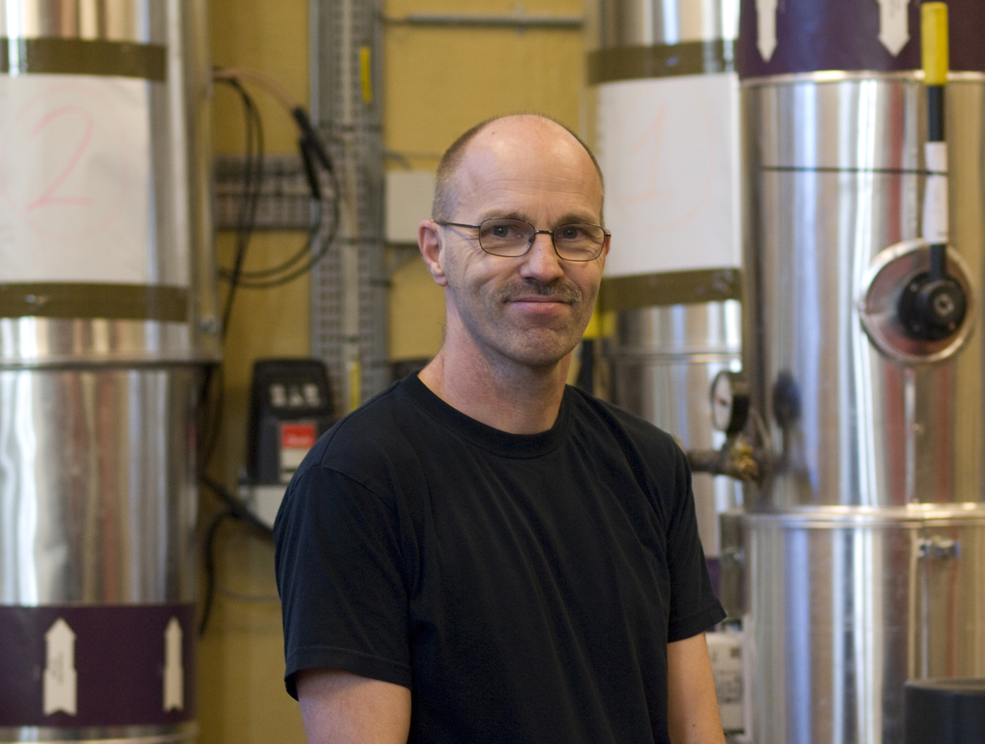 Mand med sorte briller og sort t-shirt i et industrielt værksted med store destillationsapparater i baggrunden.