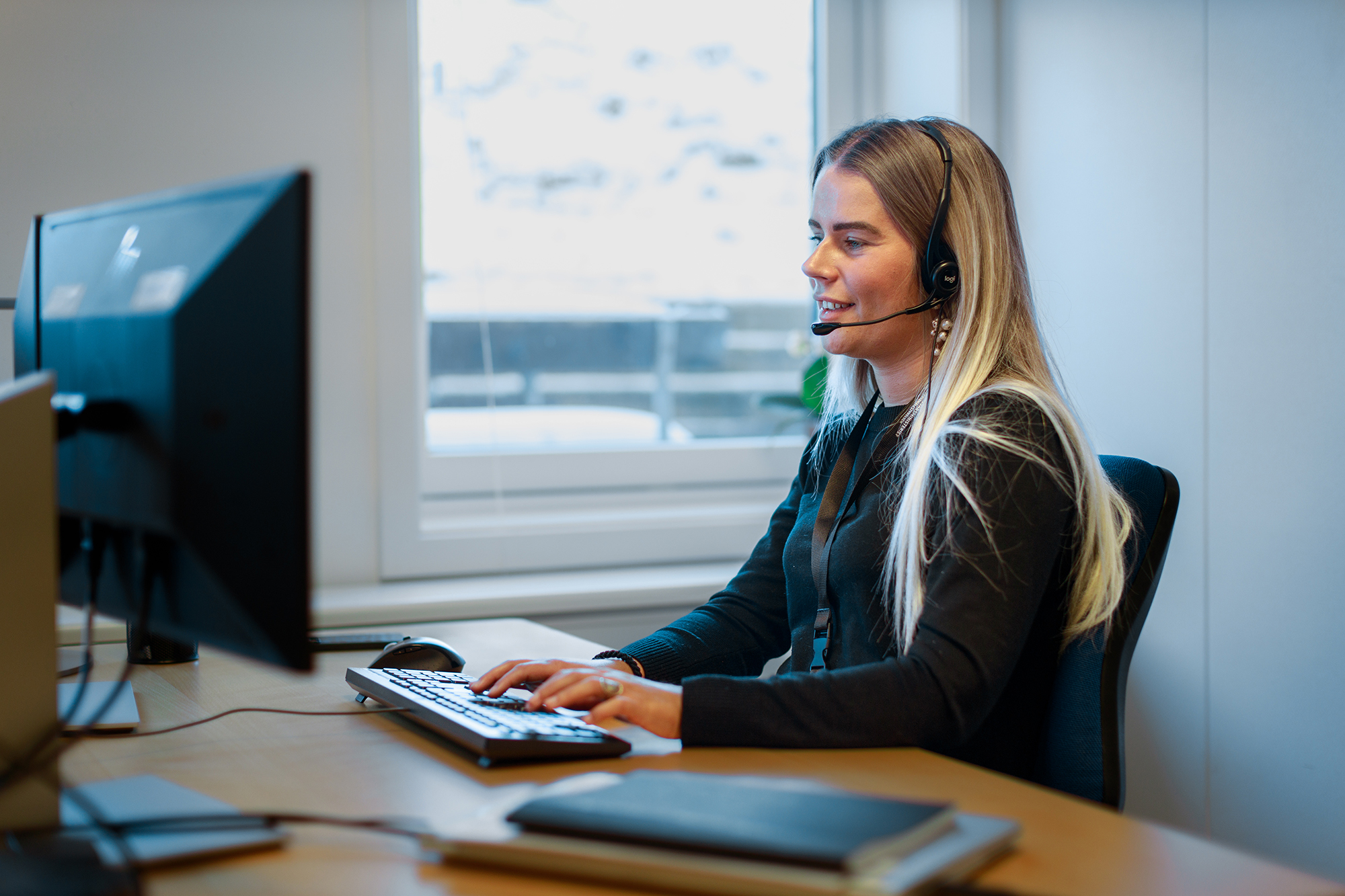 Ung kvinde med headset arbejder ved computer i kontormiljø med vinterlandskab udenfor vinduet.