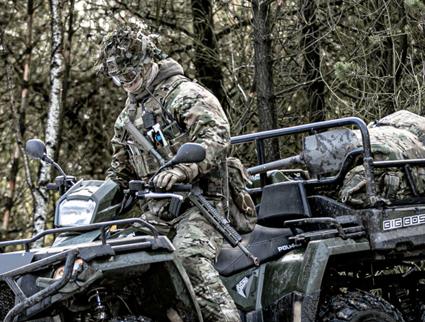 Soldat i camouflageudstyr på ATV i snedækket skov, iført maske og goggles med udstyr og riffel, klar til operation.