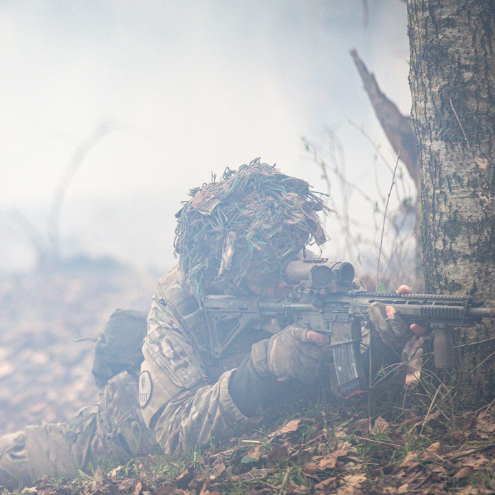 Soldat i camouflageuniform ligger skjult i tåget skov og sigter med riffel, hvilket antyder snigende militær handling.