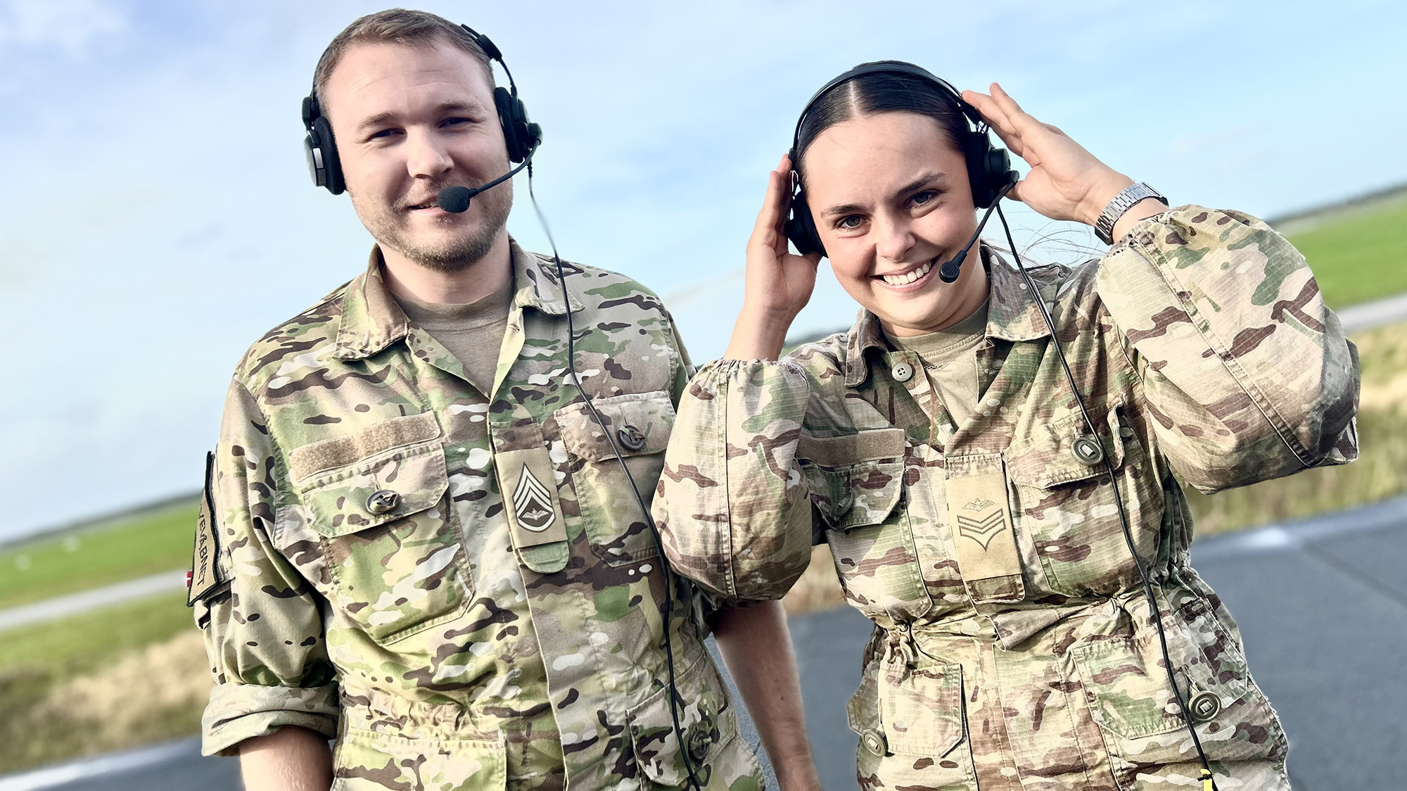 To uniformerede medarbejdere med headset står smilende udendørs ved en landingsbane.