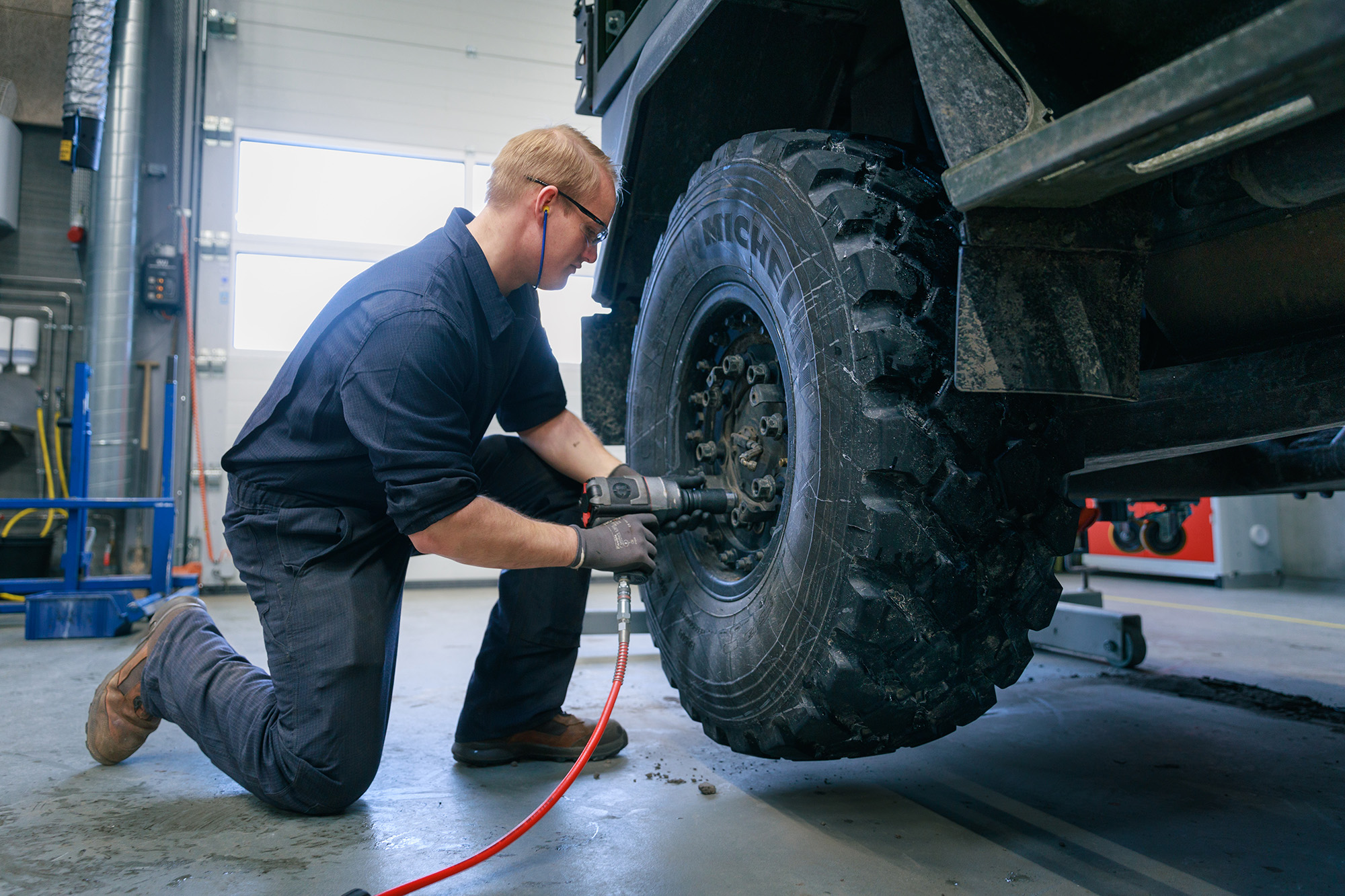 En person med sikkerhedsbriller og høreværn bruger et elværktøj til at justere et robust dæk i et lyst garageområde.