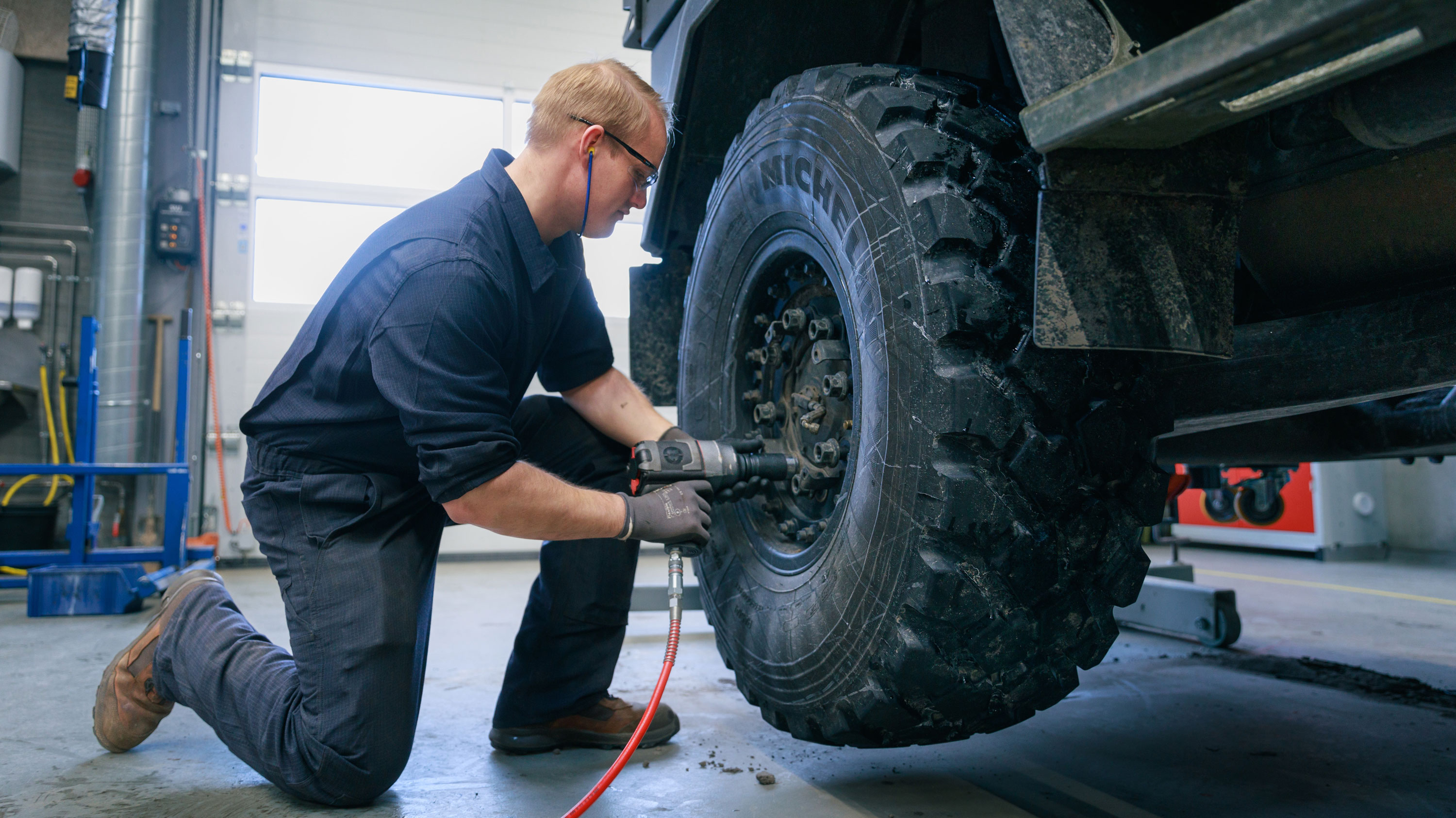 Mekaniker i mørkeblå arbejdstøj bruger trykluftsværktøj på et terrængående dæks hjul i et veludstyret værksted.