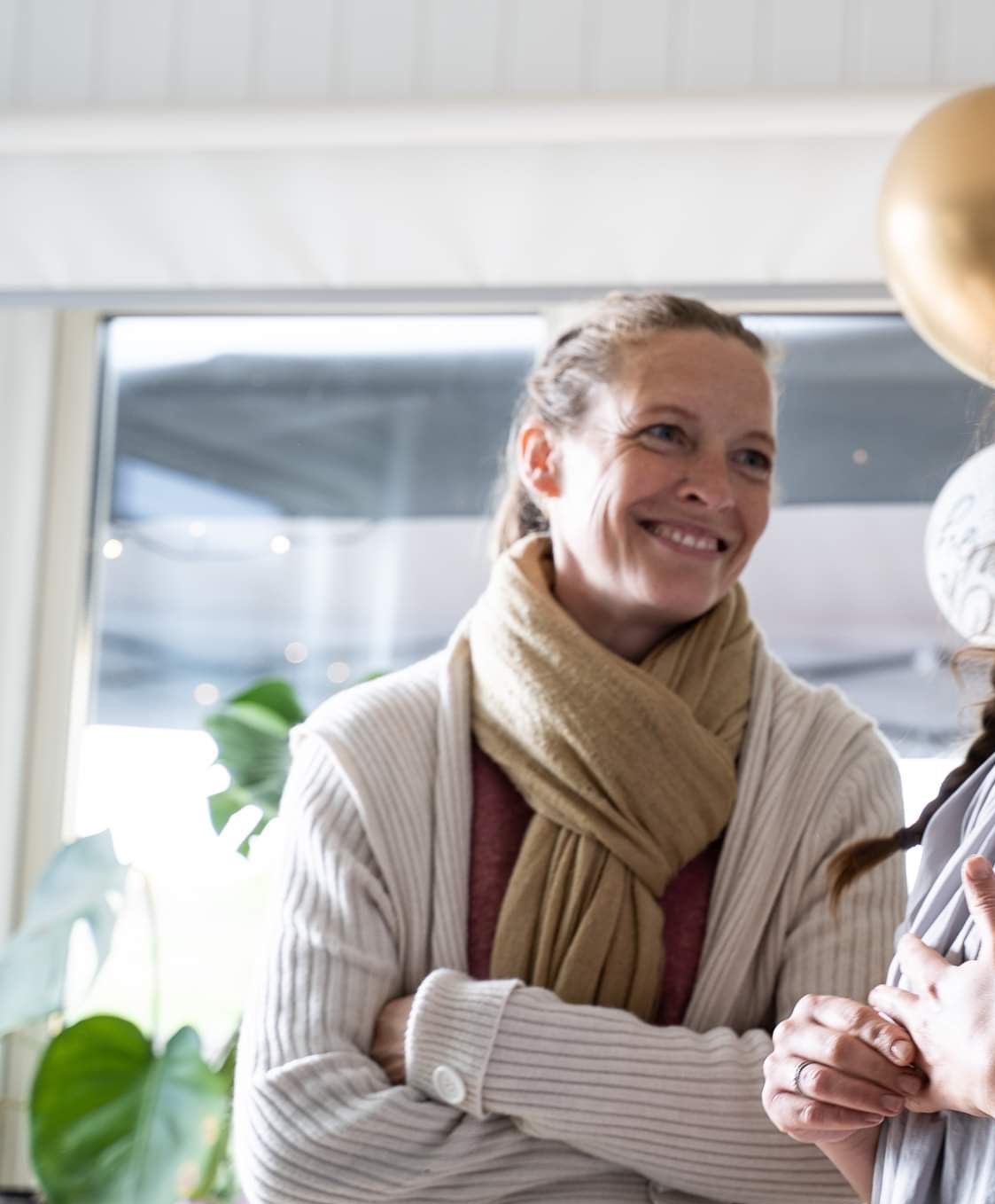 Kvinde med et varmt smil i lysefarvet cardigan og beige tørklæde står med korslagte arme og taler, omgivet af balloner og planter.