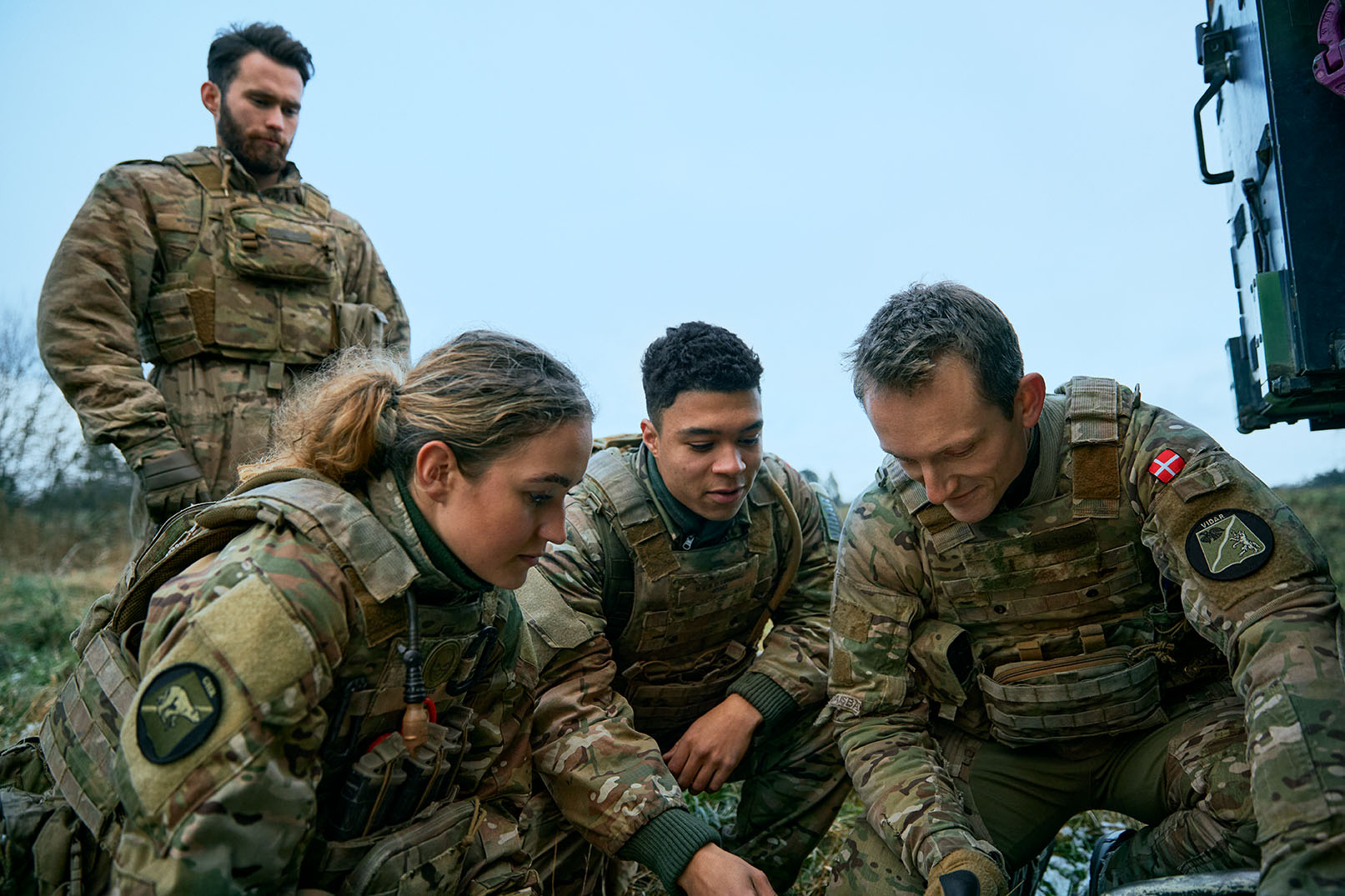 Fire soldater i camouflageuniformer arbejder sammen udendørs på græs under overskyet himmel og undersøger noget på jorden.