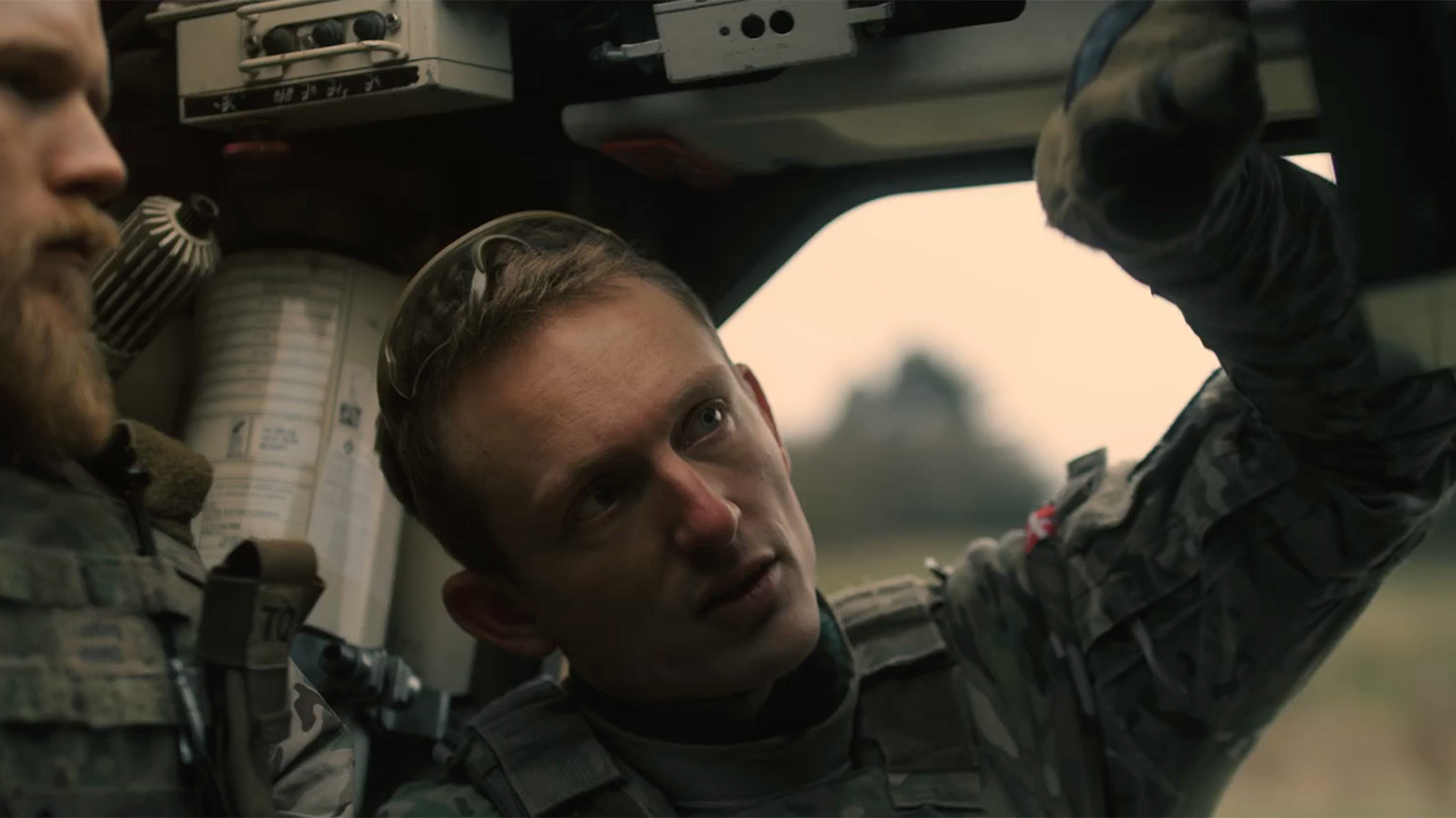 To personer i militæruniform sidder i et køretøjs kabine; en peger og taler, den anden lytter opmærksomt, omgivet af udstyr.