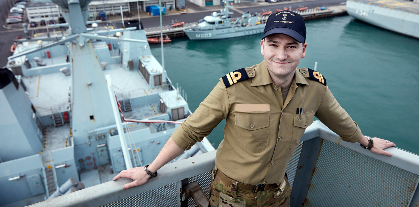 Ung mand i militæruniform med camouflagejakke og kasket med "Royal Danish Navy" sidder smilende på et skib.