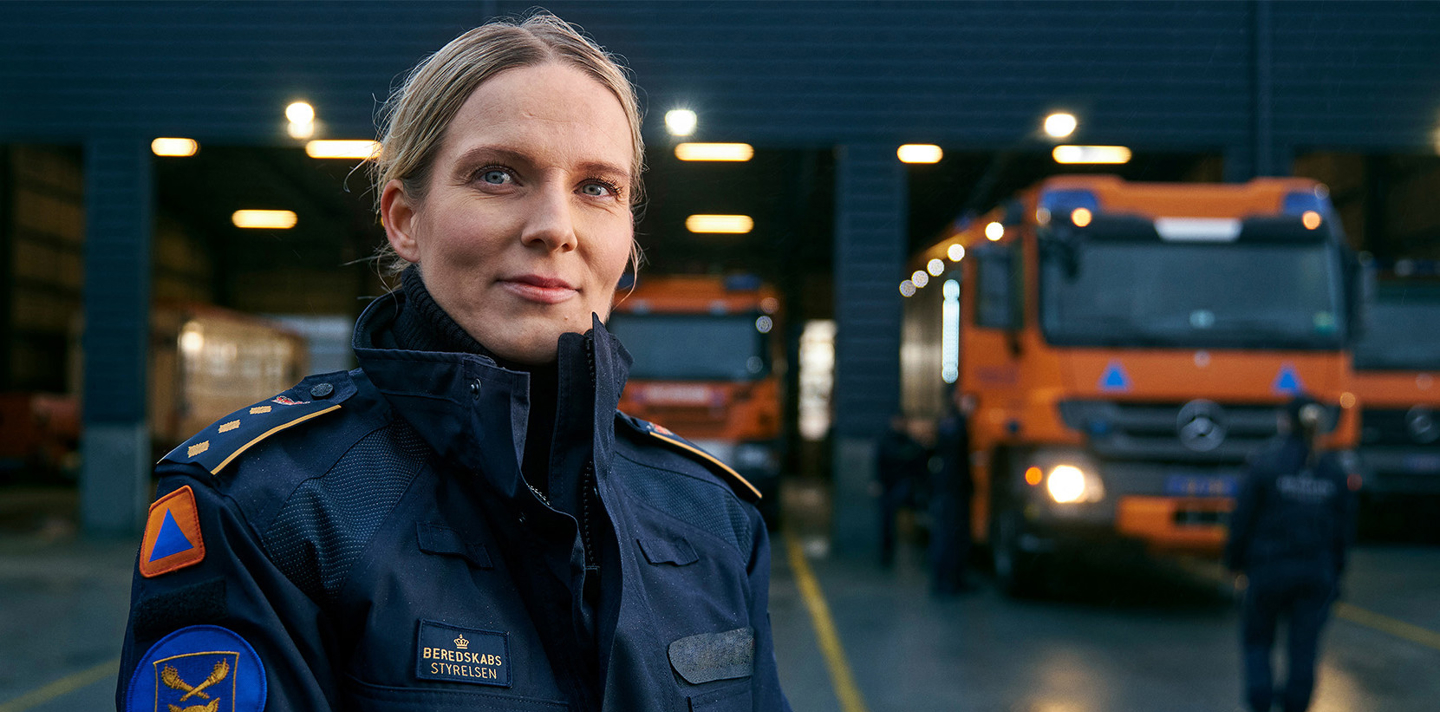 En kvinde i mørk uniform, sandsynligvis medlem af en rednings- eller beredskabsorganisation, står selvsikkert med et blåt og orange emblem synligt på uniformen. I baggrunden ses flere orange udrykningskøretøjer i en garage, hvilket understreger hendes professionelle rolle.