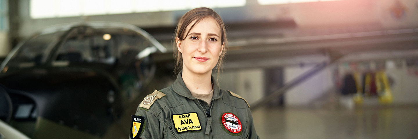 Kvinde i pilotdragt med RDAF Flying School-emblem står selvsikkert i et lyst hangar med fly i baggrunden og handsker i hånden.