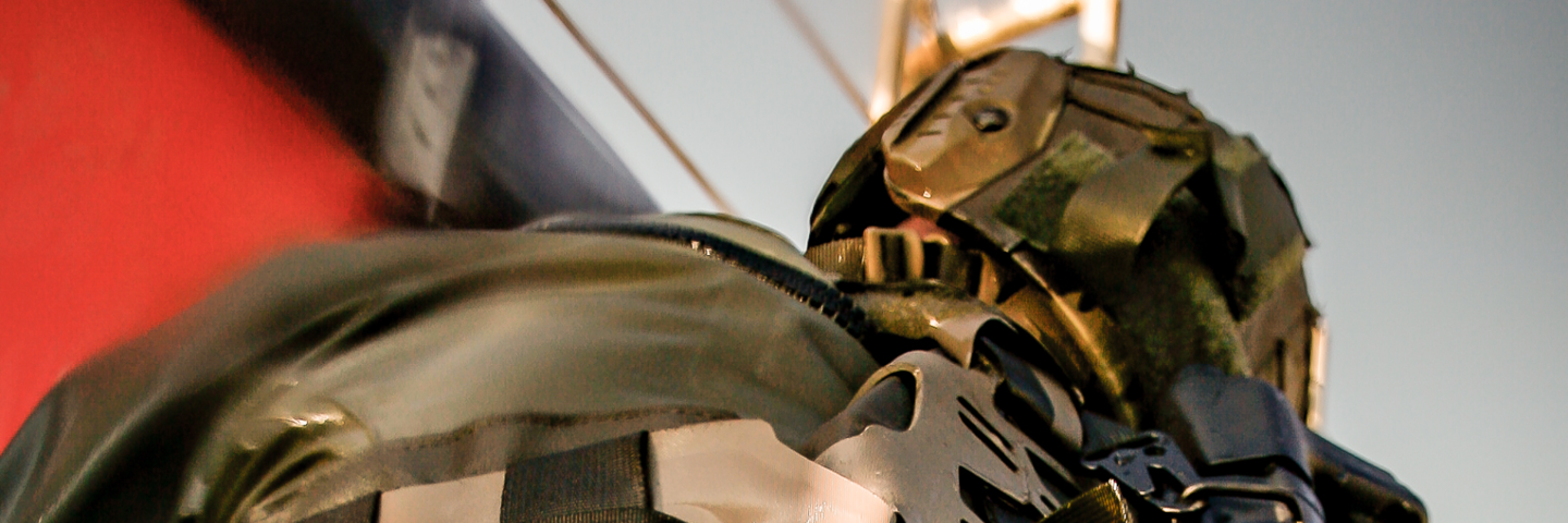 Person iført taktisk udstyr med camouflagehjelm og vest med remme og lommer i et maritimt eller industrielt miljø.