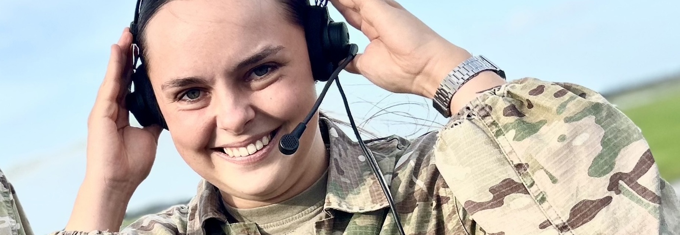 Smilende kvindelig militærperson iført camouflageuniform justerer sit headset med mikrofon, hvilket indikerer en kommunikations- eller operationsrolle; i baggrunden ses et åbent område, muligvis en flyveplads under en klar blå himmel. Hendes udtryk udstråler selvtillid og entusiasme, og detaljer som hendes ur og uniformens mønster er tydelige.
