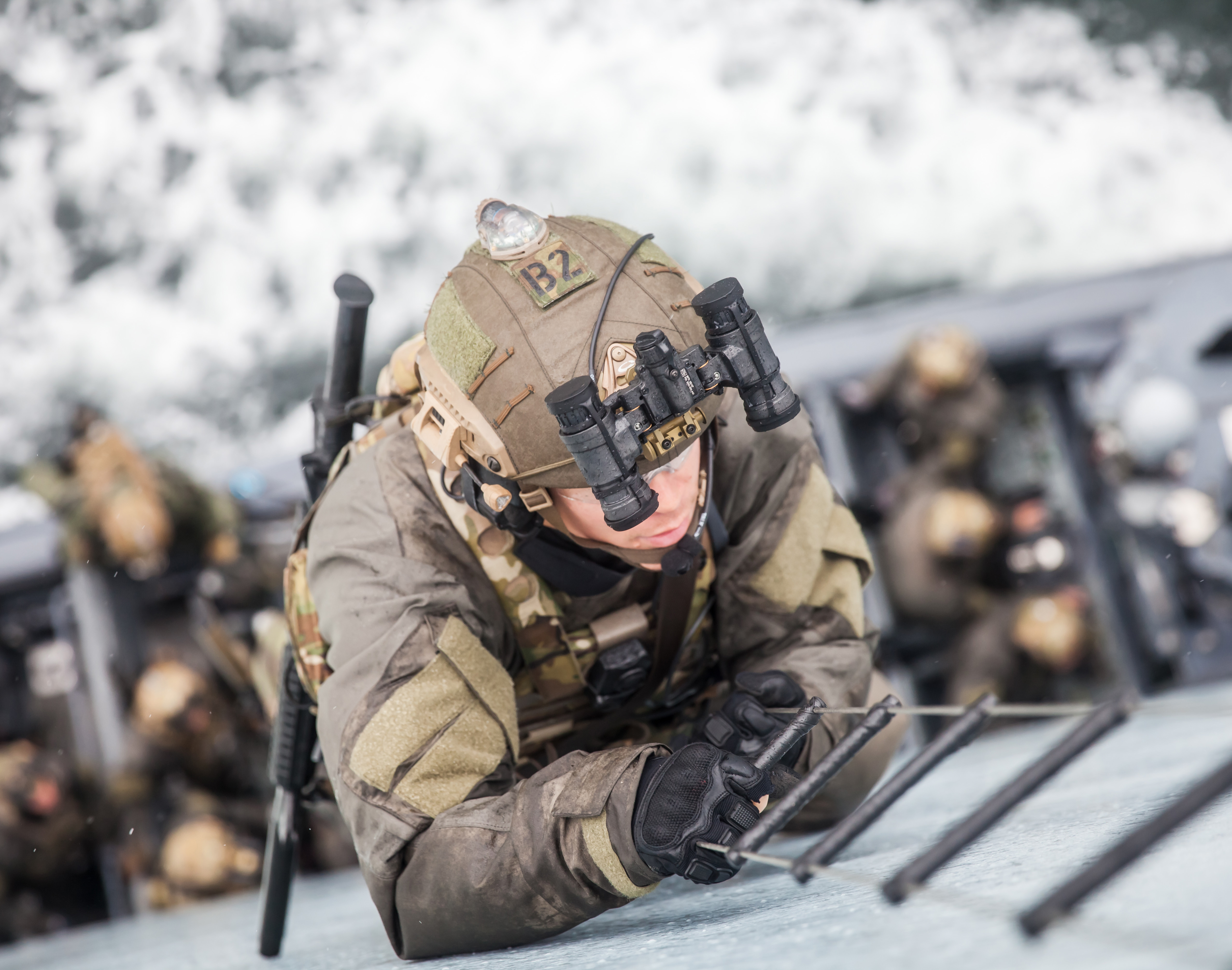 Soldat i taktisk udstyr med natkikkerter og pandelampe, liggende i maritimt miljø sammen med hold, klar til specialoperation.