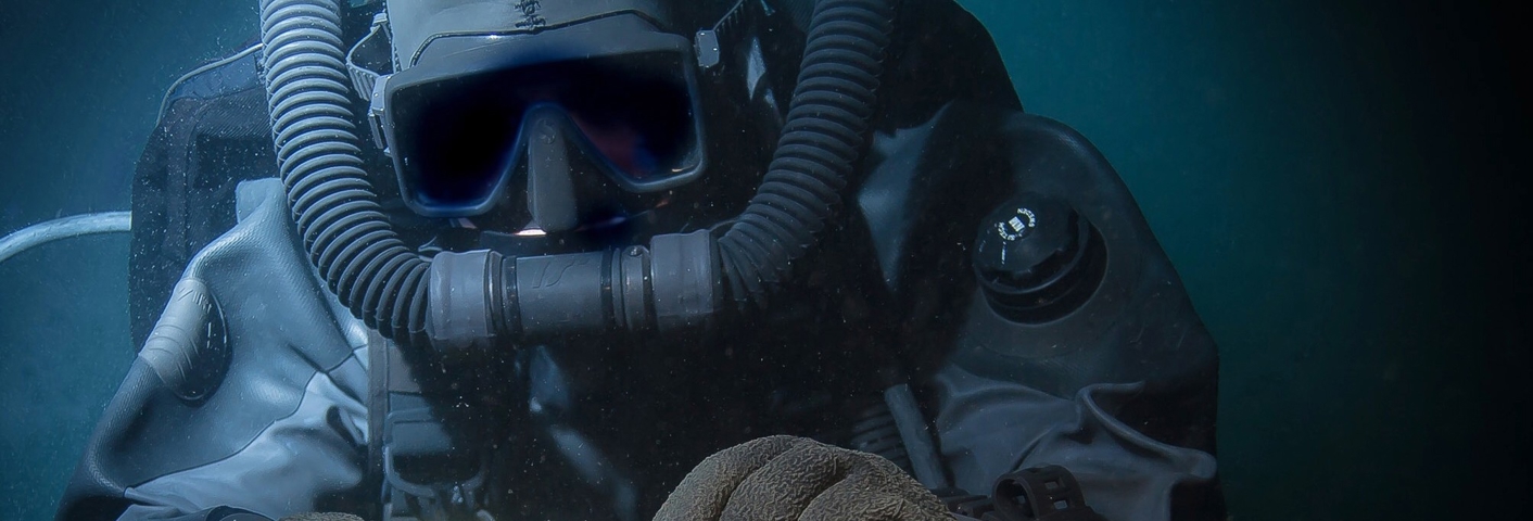 Dykker iført heldragt og fuldmaske holder kæder og reb under vandet, hvilket antyder dybhavsarbejde eller bjærgning.